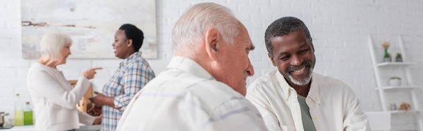 Smiling elderly interracial men talking near blurred women in nursing home, banner  - 写真・画像