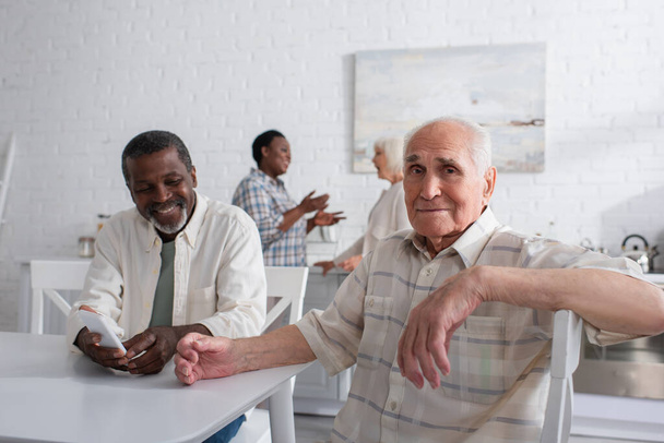 Homme âgé regardant la caméra près d'un ami afro-américain souriant avec smartphone dans une maison de retraite  - Photo, image