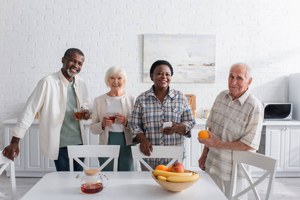 Cheerful multiethnic senior people with tea and smartphone looking at camera in nursing home  - Fotó, kép