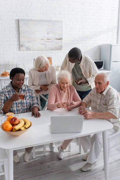 Personas mayores multiétnicas positivas que usan computadoras portátiles cerca del té y las frutas en la cocina  - Foto, Imagen