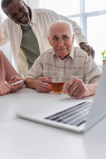 Lächelnder afrikanisch-amerikanischer Mann umarmt ältere Freunde mit Tee in der Nähe von Laptop im Pflegeheim  - Foto, Bild
