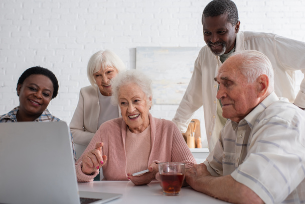 Smiling senior woman holding smartphone and pointing at laptop near interracial friends in nursing home  - Foto, Imagem