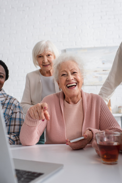Femme âgée joyeuse avec smartphone pointant vers un ordinateur portable près du thé et des amis interraciaux dans une maison de retraite  - Photo, image