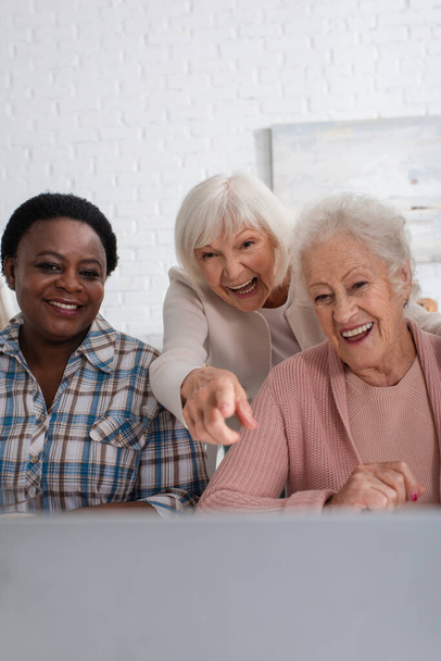 Happy senior woman pointing at laptop near interracial friends in nursing home  - Photo, image