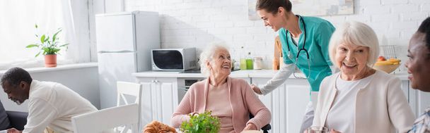Νοσοκόμα που μιλάει με ηλικιωμένες γυναίκες σε αναπηρικό καροτσάκι κοντά σε διαφυλετικούς συνταξιούχους σε γηροκομείο, πανό  - Φωτογραφία, εικόνα