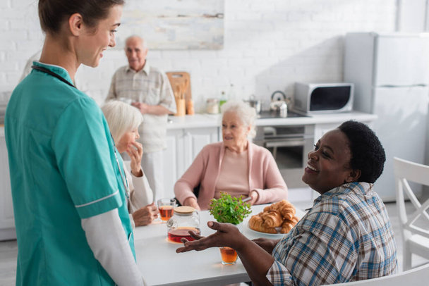 glimlachende Afrikaan amerikaanse vrouw praten met verpleegkundige in de buurt van thee en croissants in verpleeghuis  - Foto, afbeelding