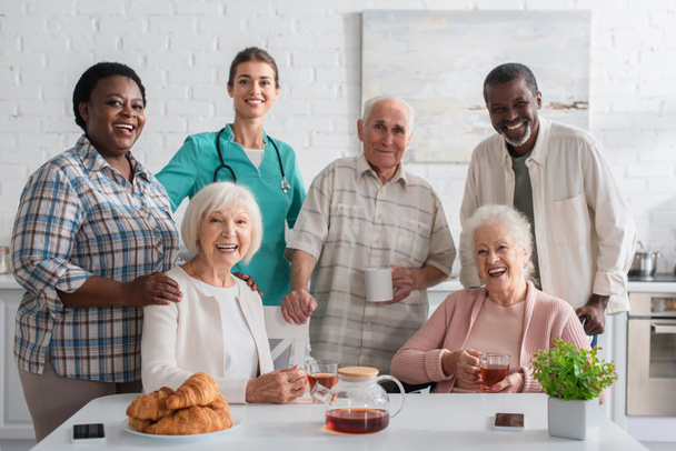 Smiling nurse standing near interracial patients and tea in nursing home  - Photo, Image
