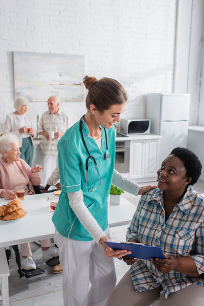 African american woman holding clipboard near smiling nurse in nursing home - Foto, Bild