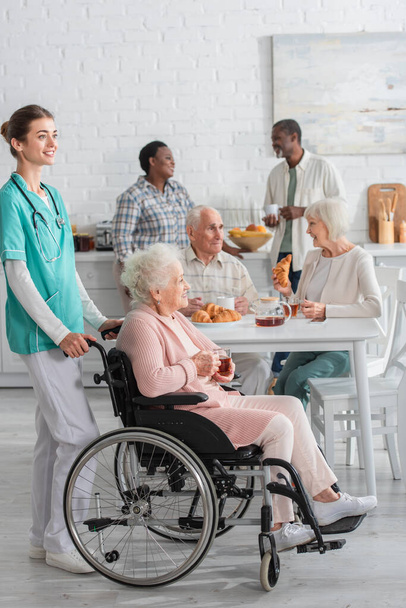 Enfermera sonriente parada cerca de una mujer mayor en silla de ruedas sosteniendo té en un hogar de ancianos  - Foto, imagen