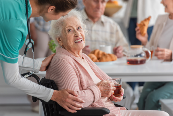 Enfermera abrazando sonriente mujer mayor con té en silla de ruedas en el hogar de ancianos  - Foto, Imagen