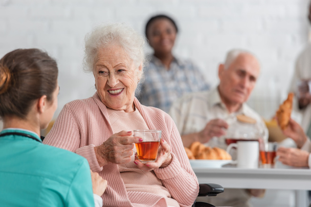 Mujer mayor positiva sosteniendo té mientras habla con la enfermera borrosa en el hogar de ancianos  - Foto, Imagen