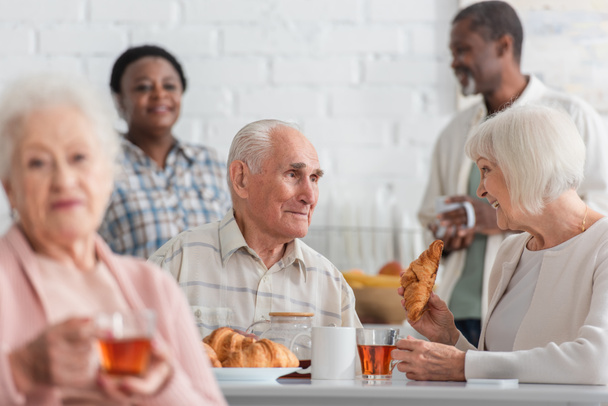 Mosolygó idős nő kezében croissant és tea közelében barátja idősek otthonában  - Fotó, kép