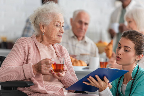 Медсестра держит планшет рядом со старшим пациентом с чаем в инвалидной коляске в доме престарелых  - Фото, изображение