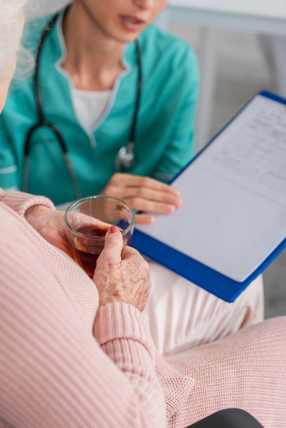 Cropped view of senior woman holding tea near blurred nurse with clipboard in nursing home  - Foto, Bild
