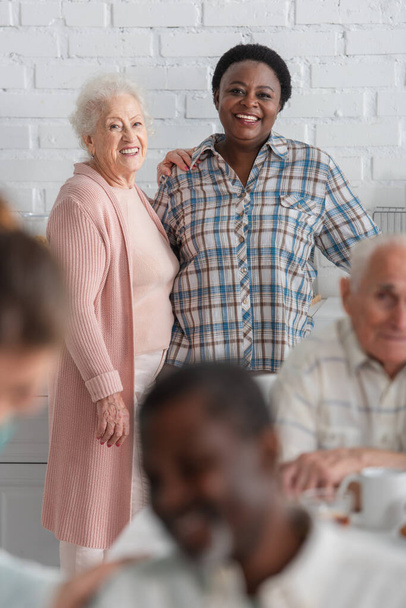 Femme souriante debout près d'un ami afro-américain dans une maison de retraite  - Photo, image