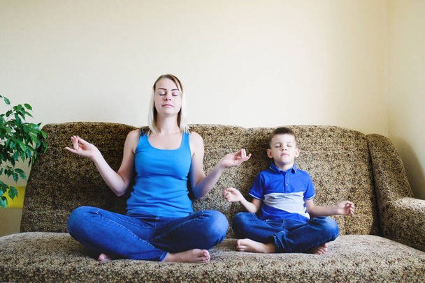 Moeder en klein kind, jongen thuis zitten op de bank, mediteren, yoga doen, ontspannen. Een gelukkige en gezonde levensstijl. Hobby en vrije tijd.  - Foto, afbeelding