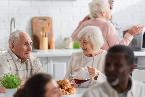 Positive elderly woman holding teapot near friend and croissants in nursing home  - Photo, Image