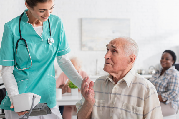 Smiling nurse holding tonometer near elderly patient in nursing home  - Zdjęcie, obraz