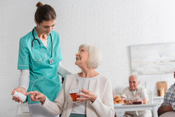 Smiling patient pointing at pills and holding tea near nurse in nursing home  - Photo, Image