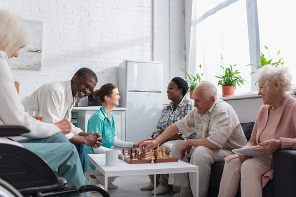 Gente interracial alegre jugando ajedrez cerca de la enfermera en un asilo de ancianos  - Foto, Imagen