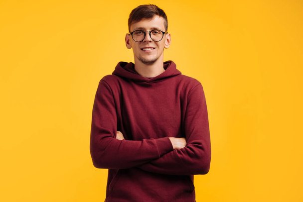Portrait of attractive man in glasses and red sweater, man standing with folded hands feels like a real leader, isolated on yellow studio background - Fotó, kép