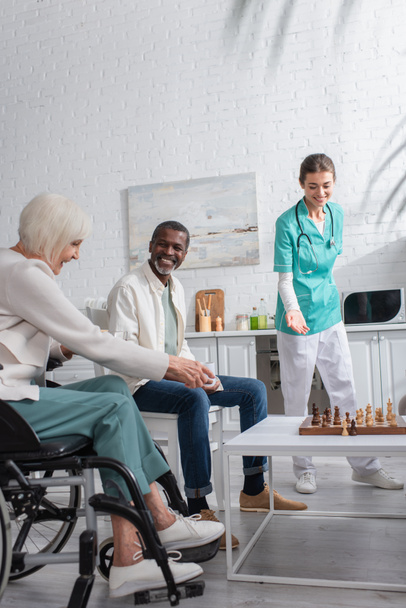 Enfermeira sorrindo apontando para tabuleiro de xadrez perto de pacientes inter-raciais em casa de repouso  - Foto, Imagem