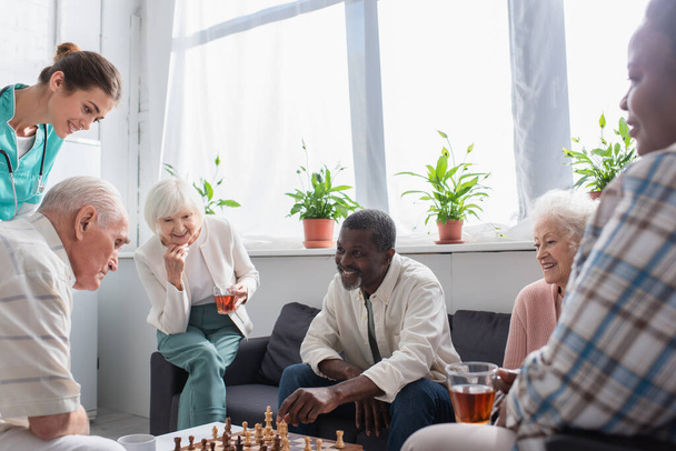 Positive interracial patients with tea playing chess near nurse in nursing home  - Foto, Imagem