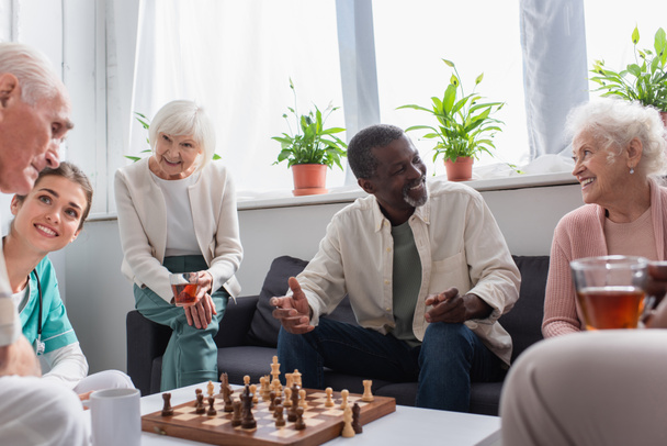 Cheerful multiethnic people with tea playing chess near nurse in nursing home  - 写真・画像