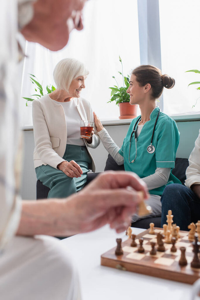 Enfermera sonriente mirando a un paciente mayor con té cerca del ajedrez en un asilo de ancianos  - Foto, Imagen