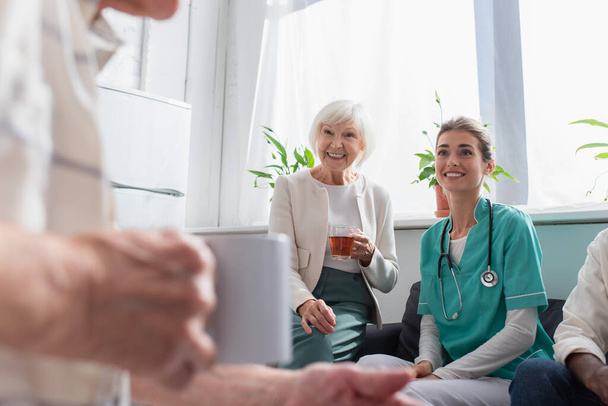 Mujer positiva con té y enfermera mirando al hombre mayor en el hogar de ancianos  - Foto, Imagen