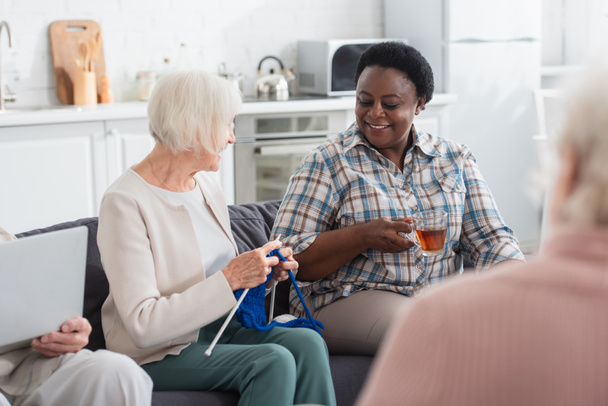 Sonrientes mujeres interracial sosteniendo té e hilo mientras habla cerca de amigo con portátil en el hogar de ancianos  - Foto, imagen