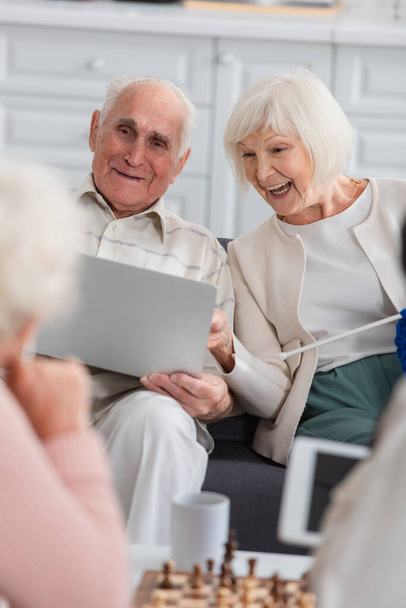 Mujer mayor positiva con hilo mirando portátil cerca de amigo y ajedrez borroso en el hogar de ancianos  - Foto, imagen