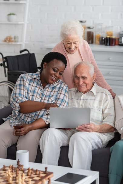 Afroamerykanka trzyma herbatę w pobliżu starszych przyjaciół z laptopem i szachami w domu opieki  - Zdjęcie, obraz