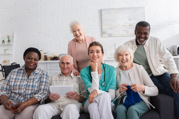 Smiling nurse sitting near interracial pensioners with yarn and digital tablet in nursing home  - Photo, Image