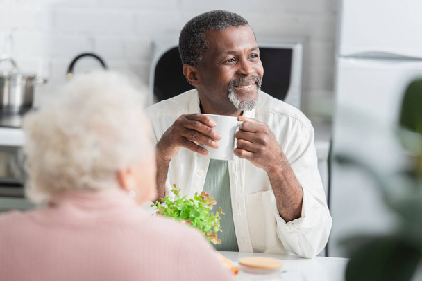 Smiling senior man holding cup near blurred friend in nursing home  - Photo, Image