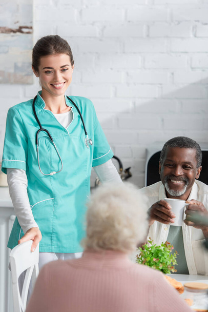 Smiling nurse standing near interracial patients in nursing home  - Фото, зображення