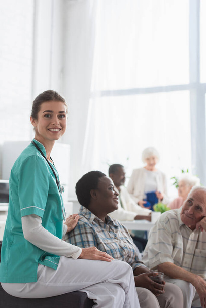 Young nurse smiling at camera near multiethnic patients in nursing home  - Photo, Image