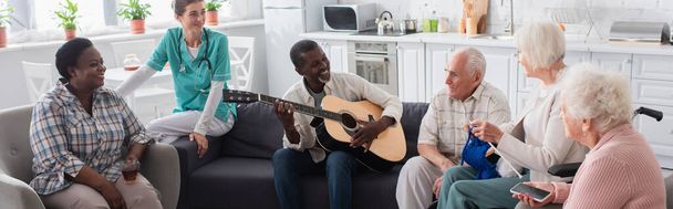 Smiling interracial elderly people playing acoustic guitar near nurse in nursing home, banner  - Foto, immagini