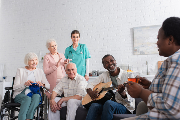 Веселые межрасовые пожилые люди с пряжей и акустической гитарой, смотрящие на африканского американского друга рядом с медсестрой в доме престарелых  - Фото, изображение