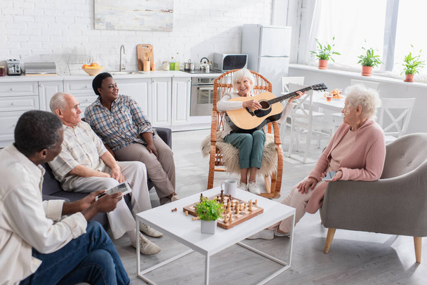 Mujer mayor alegre tocando la guitarra acústica cerca de amigos interracial y ajedrez en el hogar de ancianos  - Foto, imagen