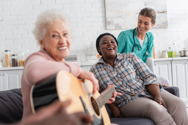 Positive interracial women with acoustic guitar looking at camera near nurse in nursing home  - Photo, image