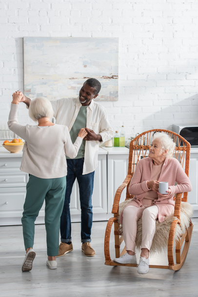 Позитивні багатонаціональні пенсіонери танцюють біля старшого друга в кріслі для камінгу в будинку престарілих
   - Фото, зображення