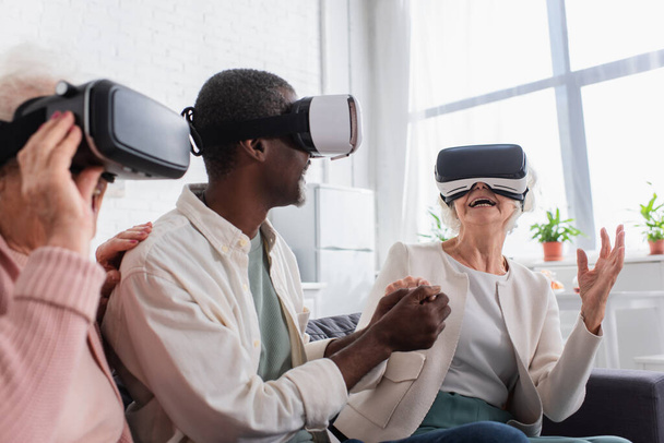 Cheerful senior woman holding hand of african american friend while gaming in vr headsets in nursing home  - Foto, Imagen