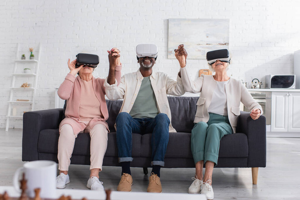Positive multiethnic pensioners holding hands while playing in vr headsets in nursing home  - Foto, imagen