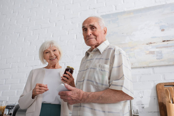 Tieffliegerblick: Senior hält Smartphone in der Nähe von Freund mit Marmeladenglas im Pflegeheim  - Foto, Bild