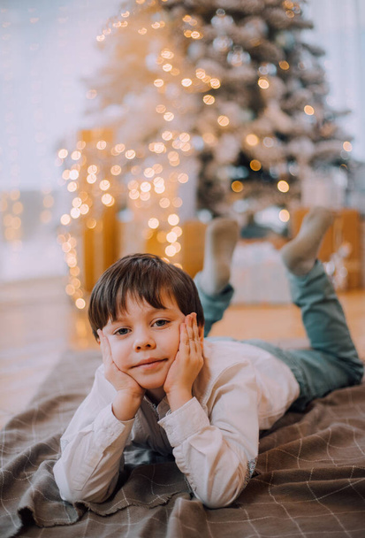 Το αγόρι βρίσκεται κάτω από τον τρόπο ζωής του δέντρου της Πρωτοχρονιάς. Πρωτοχρονιά και Χριστούγεννα. Γιορτινή διακόσμηση. - Φωτογραφία, εικόνα