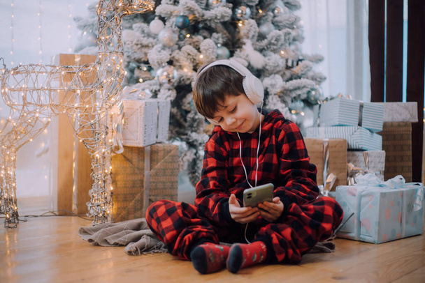 Το αγόρι με το τηλέφωνο κάτω από τον τρόπο ζωής του χριστουγεννιάτικου δέντρου. Πρωτοχρονιά και Χριστούγεννα. Γιορτινή διακόσμηση. - Φωτογραφία, εικόνα