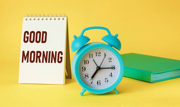 Uhr und Notizblock mit dem Text Guten Morgen auf gelbem Hintergrund, Draufsicht. - Foto, Bild