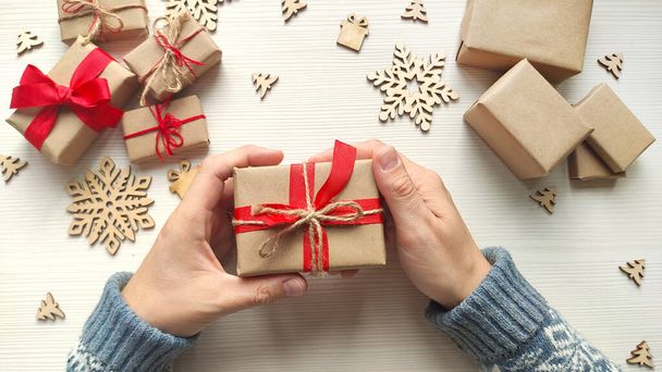 Man's hands hold christmas present in craft paper with red ribbon and jute rope. Wooden table with holiday gifts and wood snowflakes. Top view. - Foto, Imagem