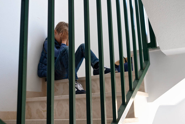 Triste chico molesto sentado en las escaleras, cubriendo su cara. Emoción negativa, depresión, estrés, concepto de bullying escolar - Foto, imagen